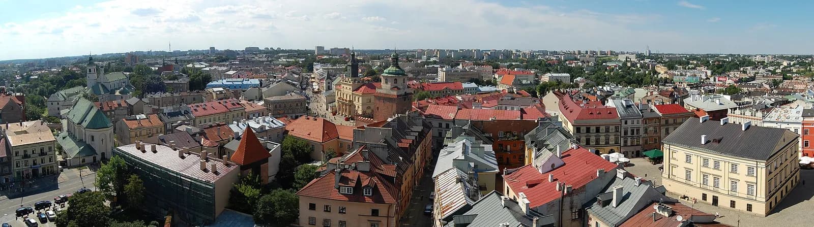 A panoramic aerial view shows various historical buildings with red and green roofs, winding streets, and a distant cityscape under a blue sky.