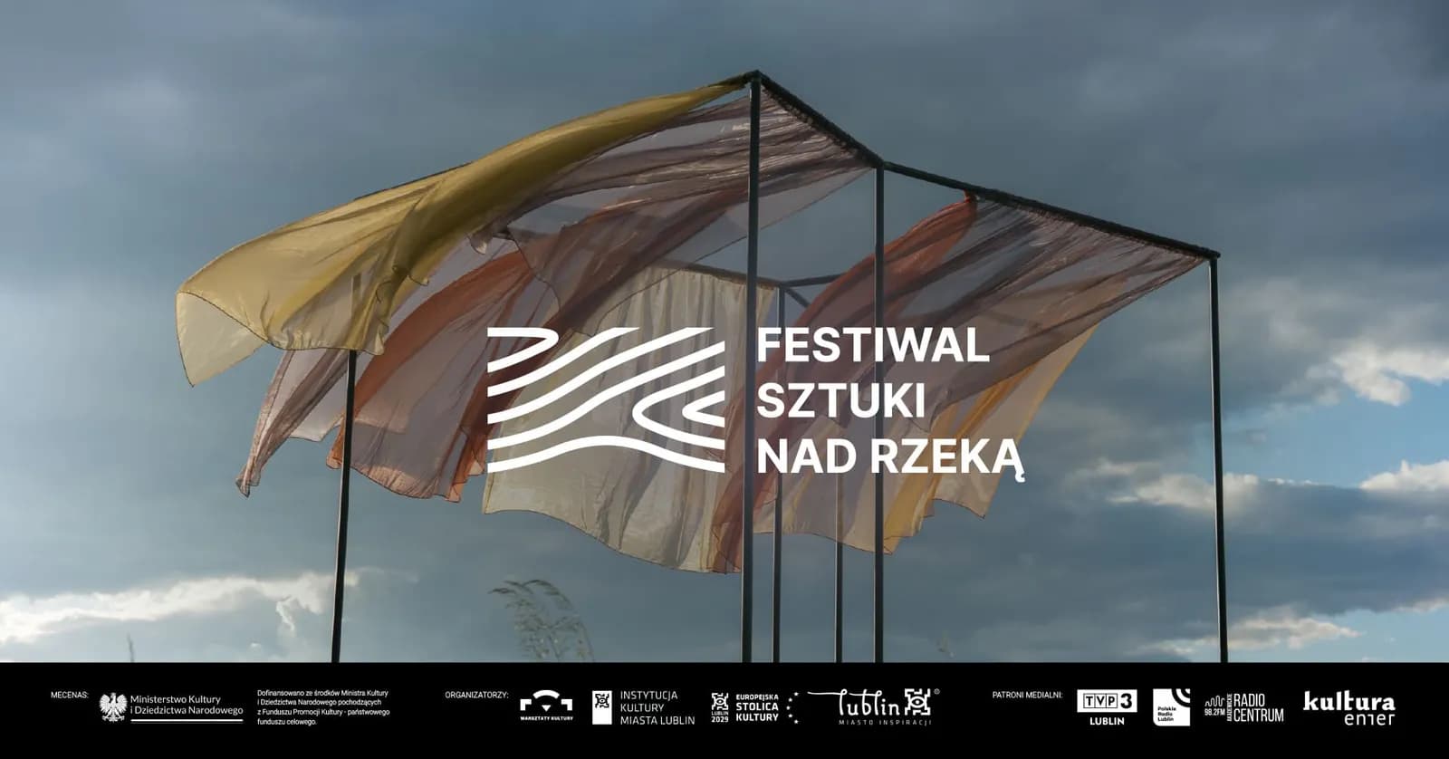 A large display with flags against a cloudy sky, with Polish text that reads "Festiwal Sztuki Nad Rzeka".